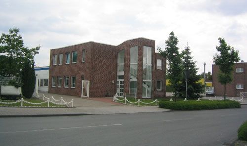Backsteingebäude mit großer Glasfront und Flachdach, umgeben von Bäumen und gepflastertem Vorplatz an einer Straße.