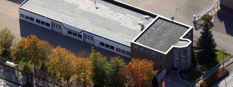 Luftaufnahme eines Flachdach-Gebäudes mit Bürotrakt aus rotem Klinker, Werkhalle und umliegenden Bäumen im Herbst.